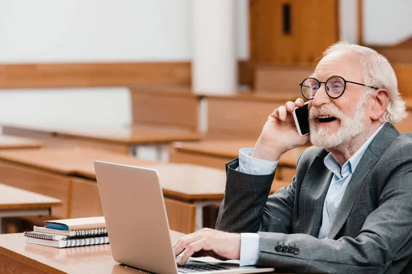 Professeur de cheveux gris souriant assis dans une salle de conférence vide et parlant par smartphone — Photo de stock