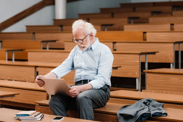 Graue Haare Professor sitzt auf Schreibtisch in leerem Hörsaal und benutzt Laptop — Stockfoto