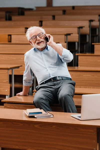 Sorridente cinza professor de cabelo sentado na mesa em sala de aula vazia e falando por smartphone — Fotografia de Stock