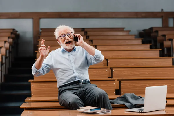 Улыбающийся седой профессор, сидящий на столе в пустой аудитории и разговаривающий по смартфону — стоковое фото