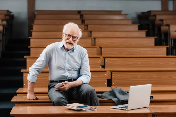 Lächelnder grauhaariger Professor sitzt im leeren Hörsaal — Stockfoto