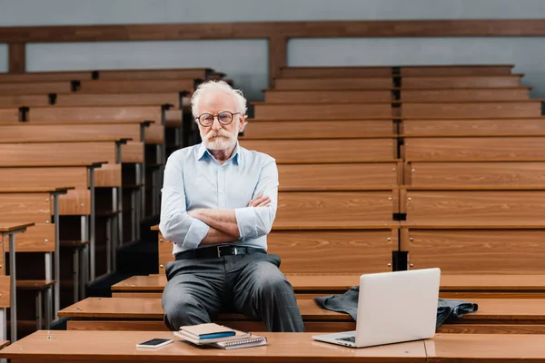 Grey hair professor sitting in empty lecture room with crossed arms — Stock Photo