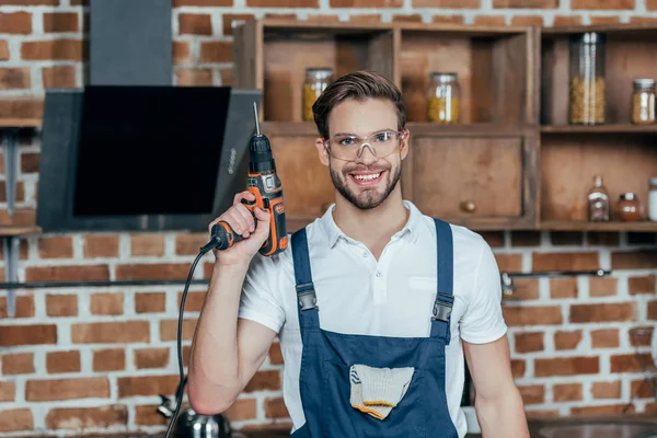 Junger Reparateur in Schutzkleidung hält Elektrobohrer in der Hand und lächelt in die Kamera — Stockfoto