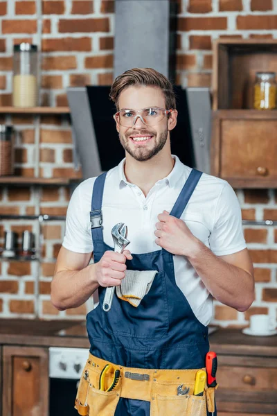 Bonito jovem reparador segurando adjustive chave inglesa e sorrindo para câmera — Fotografia de Stock