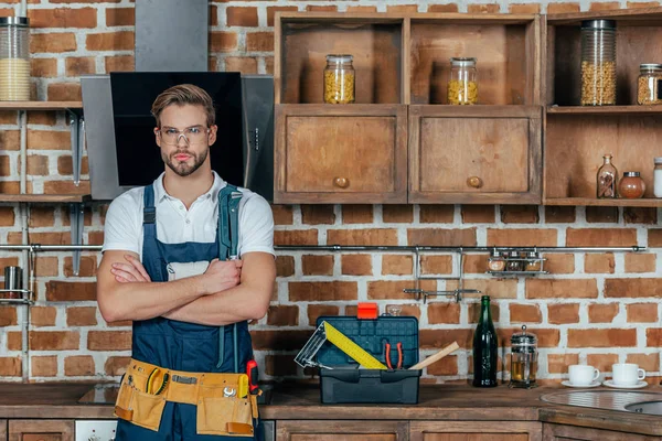 Schöner junger professioneller Werkzeugmechaniker mit Werkzeuggürtel, der mit verschränkten Armen vor der Kamera steht — Stockfoto