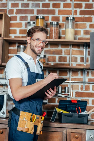 Schöner junger Vorarbeiter in Schutzkleidung, der auf Klemmbrett schreibt und in die Kamera lächelt — Stockfoto