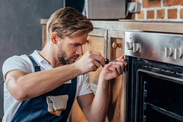 Schöner junger Reparateur, der mit einem Schraubenzieher den Ofen repariert — Stockfoto