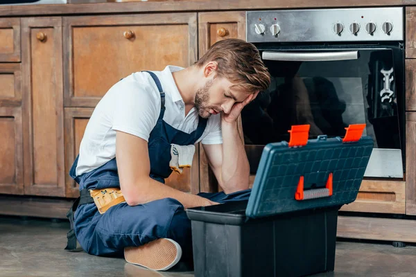 Tired young foreman sitting with toolbox near broken oven — Stock Photo