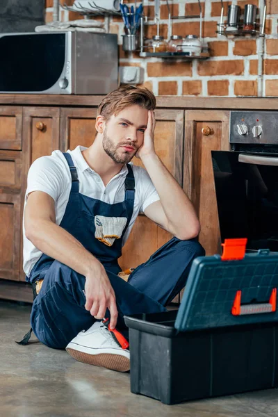 Upset young foreman sitting with toolbox near broken oven — Stock Photo
