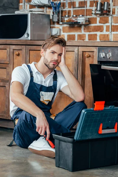 Joven capataz molesto sentado con caja de herramientas cerca del horno roto - foto de stock