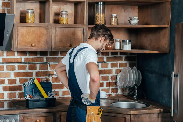 Junger Handwerker steht mit den Händen auf der Taille und blickt auf die Spüle in der Küche — Stockfoto