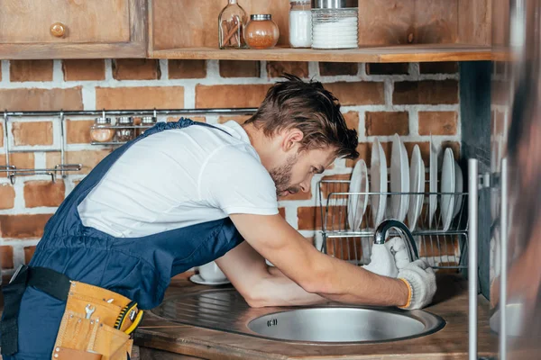 Vue latérale du jeune homme à tout faire réparant évier dans la cuisine — Photo de stock