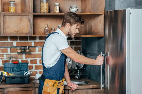 Seitenansicht eines hübschen jungen Reparateurs, der einen Kühlschrank repariert — Stockfoto