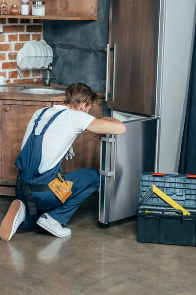 Vista trasera del reparador en ropa de trabajo protectora fijación refrigerador en la cocina - foto de stock