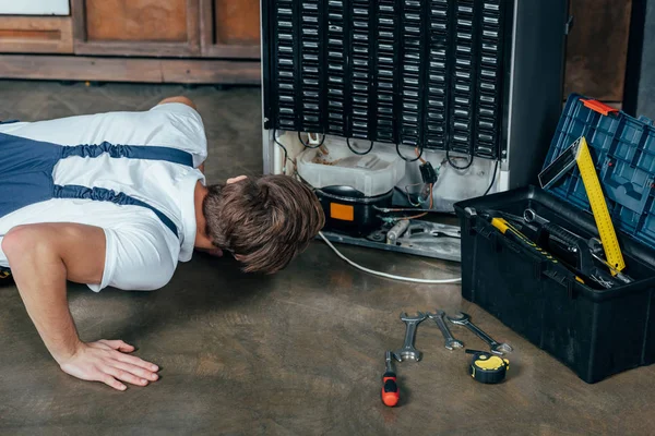 Joven reparador en ropa de trabajo protectora mirando nevera rota - foto de stock