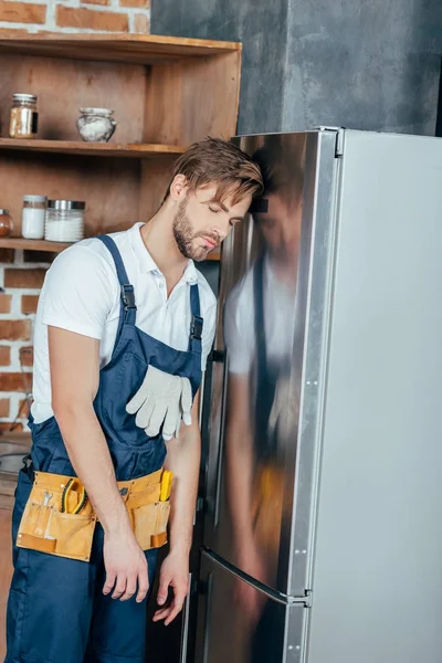 Stanco giovane caposquadra con cintura portautensili appoggiata al frigorifero rotto — Foto stock