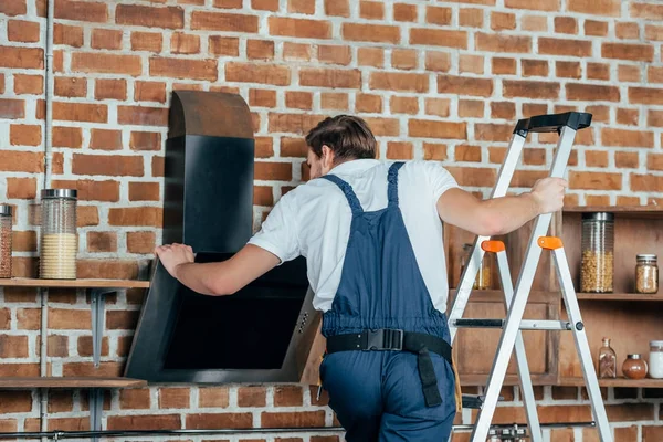 Jeune maître en vêtements de travail de protection debout sur l'échelle et la réparation hotte de cuisine — Photo de stock