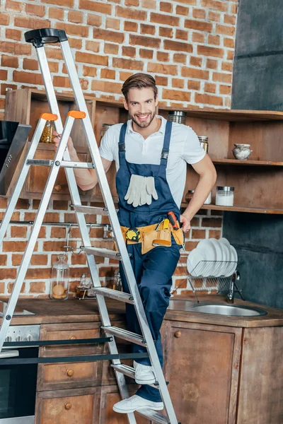 Guapo joven reparador de pie en la escalera y sonriendo a la cámara en la cocina - foto de stock