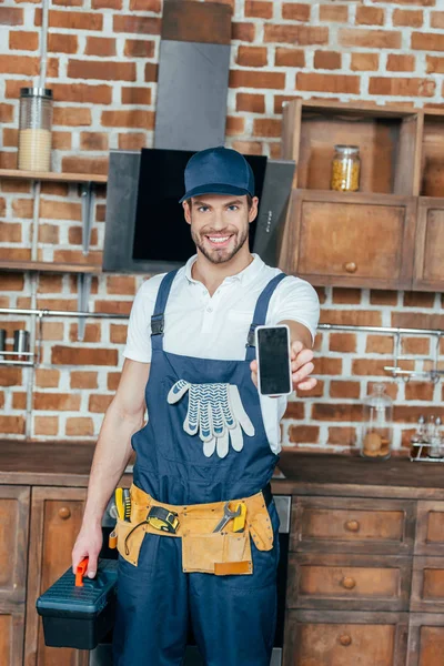 Sonriente joven hogar maestro sosteniendo teléfono inteligente y caja de herramientas - foto de stock