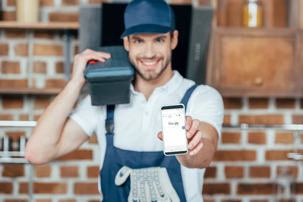 Sonriente joven hogar maestro sosteniendo teléfono inteligente con google sitio web - foto de stock