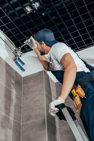 Vista de ángulo bajo del fontanero de pie en la escalera y el trabajo con tuberías en el baño - foto de stock