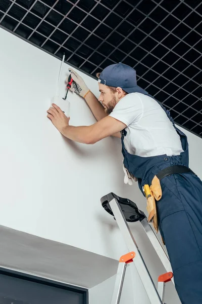 Vista basso angolo di giovane elettricista in piedi sulla scala e fissaggio allarme casa — Foto stock