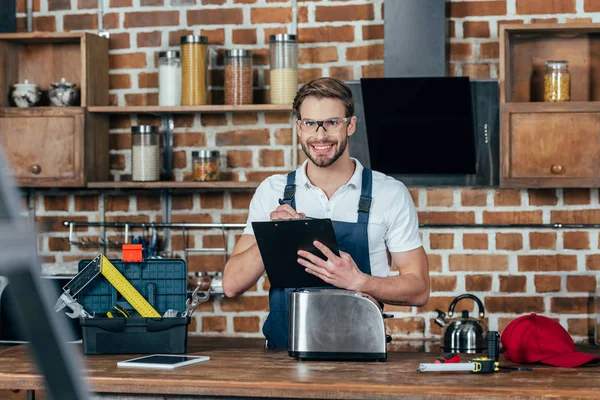 Junger Reparateur schreibt auf Klemmbrett und lächelt in die Kamera, während er Toaster repariert — Stockfoto