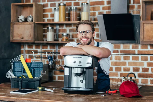 Schöner junger Reparateur lehnt an Kaffeemaschine und lächelt in die Kamera — Stockfoto