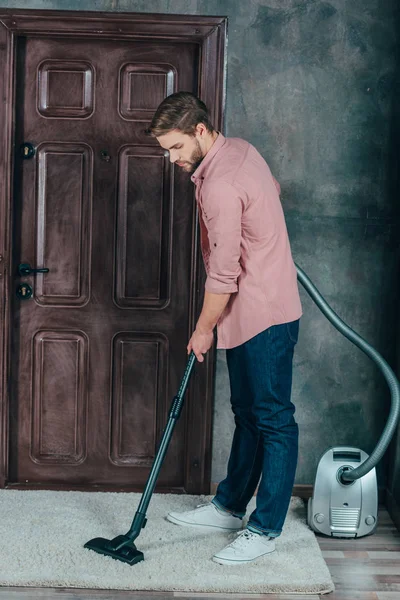 Junger Mann benutzt Staubsauger und putzt Teppich zu Hause — Stockfoto