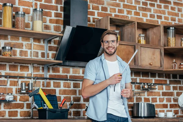 Молодий чоловік тримає вимірювальну стрічку і посміхається на камеру під час ремонту кухонної витяжки — стокове фото