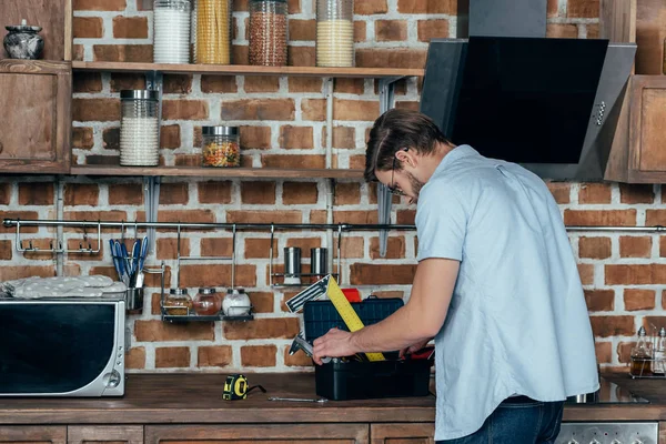 Junger Mann sucht in Werkzeugkiste mit Werkzeug, während er in Küche arbeitet — Stockfoto