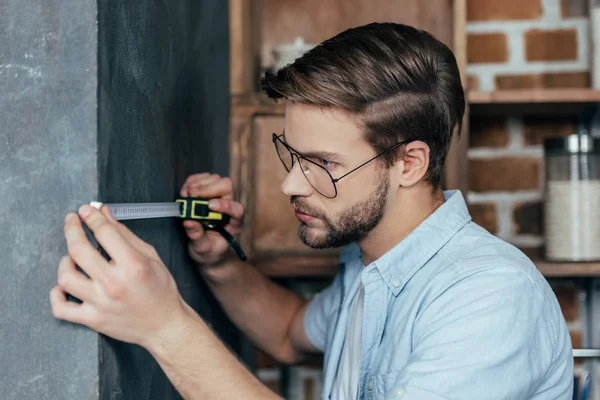 Jeune homme en lunettes mesurant mur avec du ruban adhésif à la maison — Photo de stock