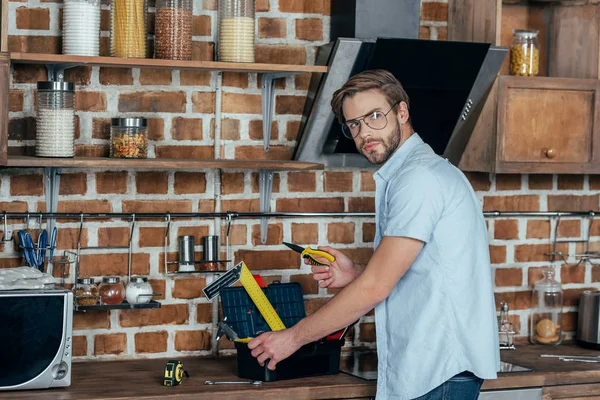 Молодой человек в очках берет инструменты из ящика с инструментами и смотрит на камеру на кухне — стоковое фото