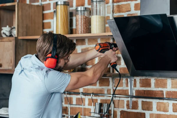 Junger Mann in Ohrenschützern bohrt Küchenhaube mit Elektrobohrmaschine an — Stockfoto