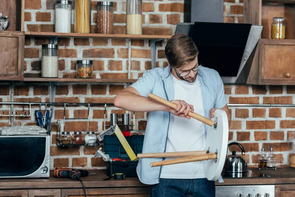 Концентрований молодий чоловік в окулярах ремонтує табурет на кухні — стокове фото