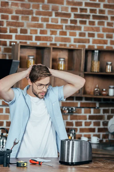 Giovane uomo in occhiali che si tiene per mano dietro la testa mentre ripara tostapane a casa — Foto stock