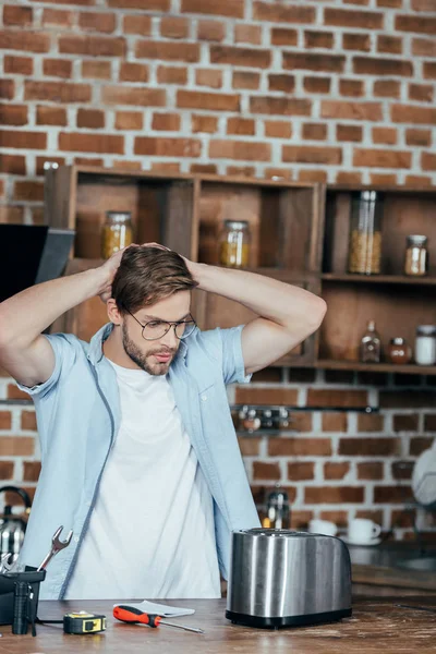 Jeune homme à lunettes tenant la main derrière la tête tout en réparant grille-pain à la maison — Photo de stock