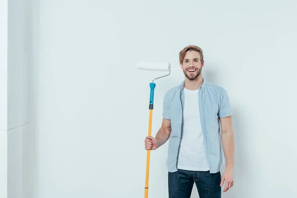 Jovem bonito segurando rolo de pintura e sorrindo para a câmera em cinza — Fotografia de Stock