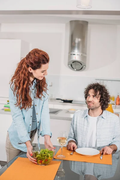 Sorrindo namorada colocando salada na mesa e olhando para o namorado — Fotografia de Stock