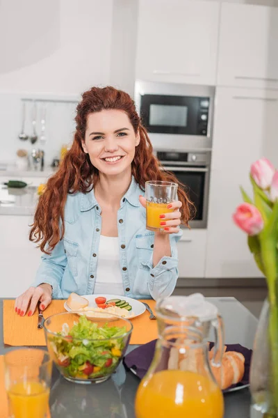 Menina sorridente sentada à mesa com refeição e segurando um copo de suco de laranja — Fotografia de Stock
