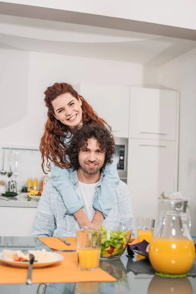 Happy girlfriend hugging boyfriend from back at kitchen and they looking at camera — Stock Photo