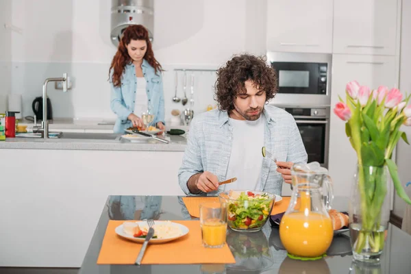 Novio comer mientras novia cocinar en la cocina - foto de stock
