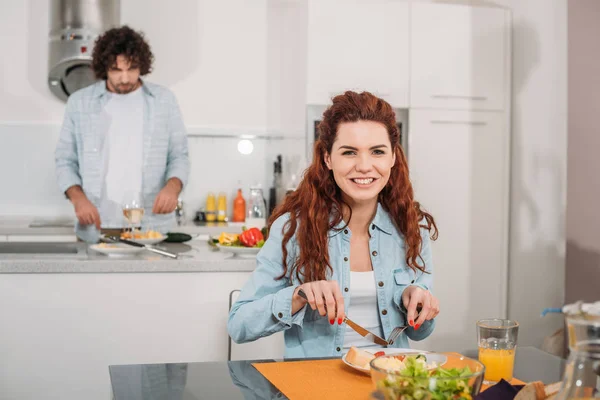 Smiling girlfriend eating while boyfriend cooking at kitchen — Stock Photo