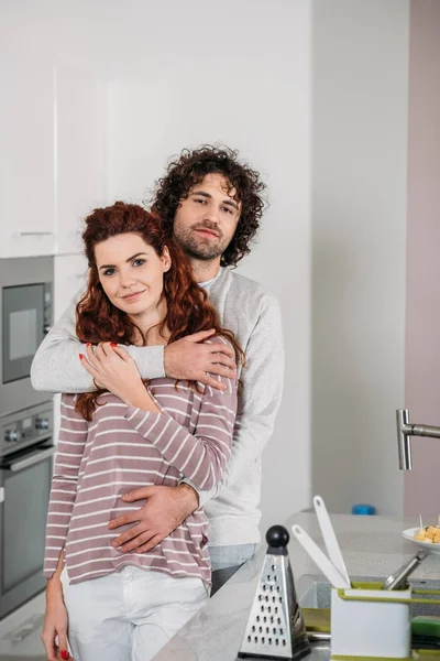 Namorado abraçando namorada na cozinha e eles olhando para a câmera — Fotografia de Stock