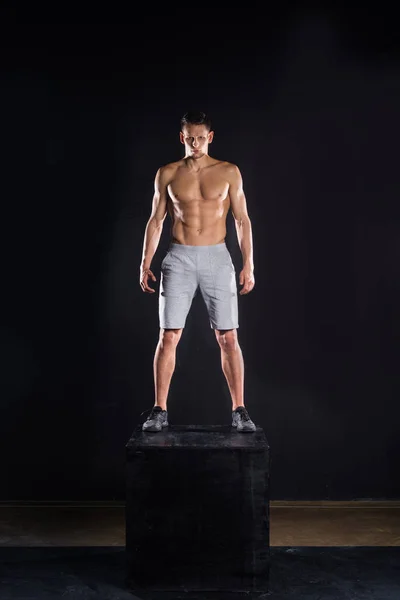 Sportivo muscoloso senza maglietta in piedi sul blocco e guardando la fotocamera su nero — Foto stock