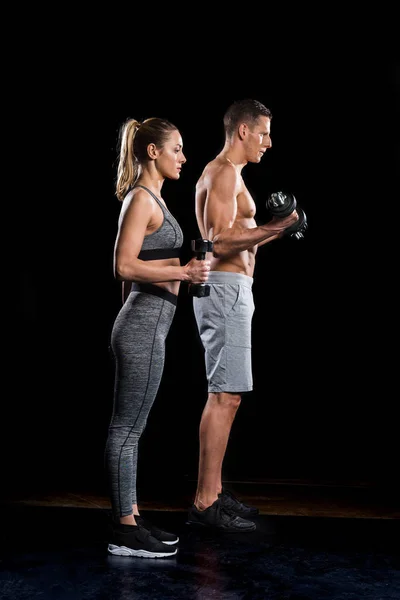 Vue latérale du jeune couple sportif faisant de l'exercice avec des haltères sur noir — Photo de stock