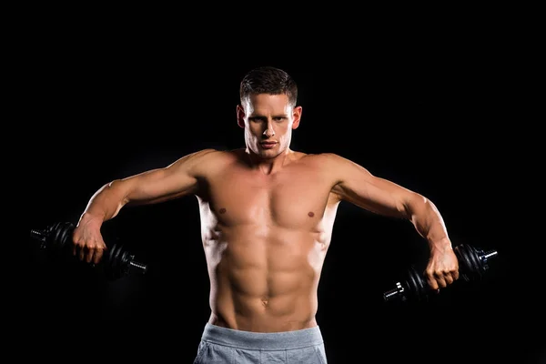 Young muscular shirtless sportsman holding dumbbells and looking at camera isolated on black — Stock Photo