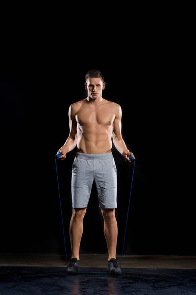 Handsome young sportsman standing with skipping rope and looking at camera on black — Stock Photo