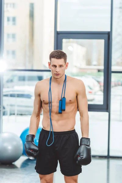 Junger muskulöser Sportler in Boxhandschuhen mit Springseil blickt in Turnhalle in die Kamera — Stockfoto