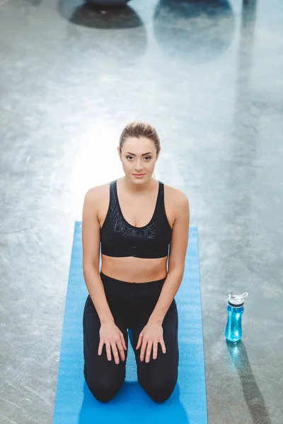Vue grand angle de la jeune sportive assise sur un tapis de yoga et regardant la caméra dans un studio de fitness — Photo de stock